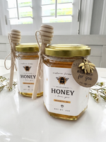 Two jars of honey with gold lids on a white surface, featuring a bee-themed label and tag.