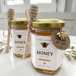 Two jars of honey with gold lids on a white surface, featuring a bee-themed label and tag.